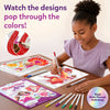 A young girl seated at a wooden table smiling while coloring Paint-Over Art sheets with markers; completed bright floral and fantasy designs visible, open kit box and scattered markers around, text reads Watch the designs pop through the colors, Magic Shine-Through badge shown.