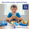 Young boy smiling while holding an educational card from the Skillmatics Learn About Real Things set, seated at a table with the open box, illustrated concept cards, and storage tray beside him. Text reads “Real-world concepts, explained simply” and “Ages 7–14,” showing hands-on learning with colorful cards designed to teach everyday topics.