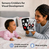Lifestyle image for Skillmatics Sensory Crinklers. A mother smiles holding two cloth crinklers in front of a baby on a bed in pink outfit. Baby looks at star, butterfly and bee patterns. Text reads “Sensory Crinklers for Visual Development” with crinkle sound and wear and play icons.
