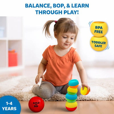 A toddler sits on a soft rug indoors, balancing colourful wobbly pieces from the Build And Bop toy while a red ball labeled “Build And Bop” sits nearby. Text at the top reads “Balance, bop, & learn through play!”. Two yellow icons on the right state “BPA Free” and “Toddler Safe,” and a blue badge on the bottom left indicates the toy is for ages 1–4 years.