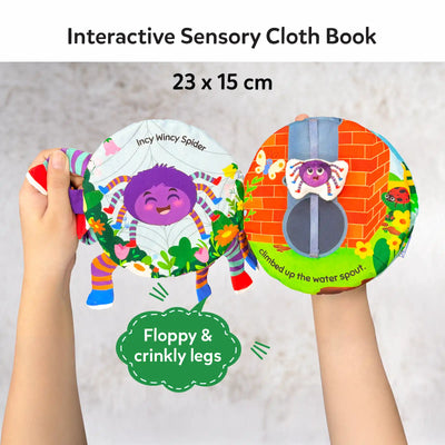 Hands holding an open Incy Wincy Spider interactive sensory cloth book. The left page shows a cheerful purple spider illustration with colorful legs and flowers, while the right page features a brick wall scene with a fabric spider that moves along a vertical strap. A callout highlights floppy and crinkly legs. The soft fabric pages, stitched details, and movable elements encourage tactile play and exploration.