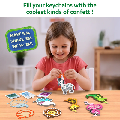 A young child sits at a wooden table smiling while holding a zebra-shaped foam shake-it keychain filled with confetti. On the table are multiple animal foam shapes, transparent covers, small confetti packets, and finished keychains in bright colors. A text callout reads “Make ’em, shake ’em, wear ’em,” with a softly blurred indoor background and natural lighting.