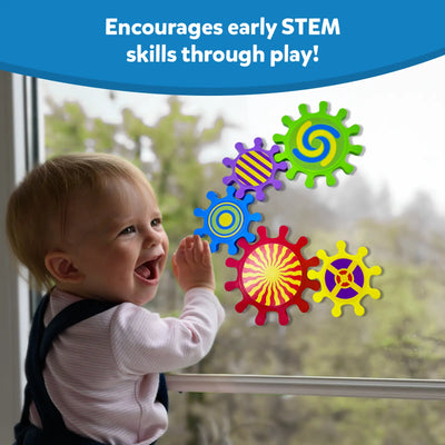 A laughing baby in a pink top and denim overalls reaches up to touch five Skillmatics Spinny Gears stuck to a large window. Gears display vivid patterns: green spiral, purple striped, blue concentric, red sunburst, and yellow cross. Trees are visible outside. Header reads "Encourages early STEM skills through play!"