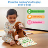 A smiling baby in printed pajamas sits on the floor gently pressing the tail of a brown plush monkey toy. The monkey holds a polka-dotted cloth printed with “Peek-a-boo, where are you?” At the top, blue text reads “Press the monkey’s tail to play peek-a-boo!” Icons on the right show “Battery Operated,” “Soft & Fuzzy,” and “Do Not Wash” in green, orange, and pink labels.
