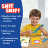A young child sitting at a table smiles while using green safety scissors to cut a blue paper strip from the Snip Snip activity set. Open activity pages with Disney characters lie on the table alongside a glue stick and an extra pair of scissors. A blue panel lists features such as developing scissor skills, mess-free play, step-by-step learning, and creative activities with Mickey and friends.