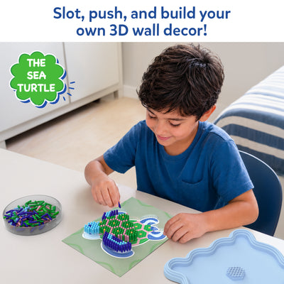 Young child seated at a table creating a 3D sea turtle artwork with the Skillmatics Stixels kit. The child places colorful Stixels into a transparent turtle-shaped base using the color guide, while loose Stixels and the decorative display frame are arranged nearby in a bright indoor setting.