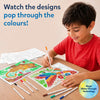 Child sitting at table smiling while using paint-over art markers on illustrated activity sheets, revealing bright colors through hidden patterns; multiple markers laid out on wooden surface and finished colorful animal designs visible beside ongoing coloring activity.
