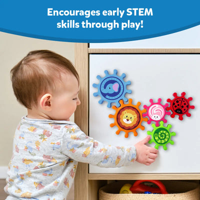 A toddler in a grey floral top reaches to touch Skillmatics Spinny Gears mounted on a white cabinet door. Five interlocking animal gears are visible: blue elephant, orange lion, pink rabbit, red ladybug, and green caterpillar. A toy basket sits below the cabinet. Header reads "Encourages early STEM skills through play!"