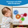 A toddler in a grey floral top reaches to touch Skillmatics Spinny Gears mounted on a white cabinet door. Five interlocking animal gears are visible: blue elephant, orange lion, pink rabbit, red ladybug, and green caterpillar. A toy basket sits below the cabinet. Header reads 