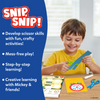 A young child sitting at a table smiles while using green safety scissors to cut a blue paper strip from the Snip Snip activity set. Open activity pages with Disney characters lie on the table alongside a glue stick and an extra pair of scissors. A blue panel lists features such as developing scissor skills, mess-free play, step-by-step learning, and creative activities with Mickey and friends.
