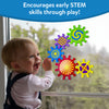 A laughing baby in a pink top and denim overalls reaches up to touch five Skillmatics Spinny Gears stuck to a large window. Gears display vivid patterns: green spiral, purple striped, blue concentric, red sunburst, and yellow cross. Trees are visible outside. Header reads 