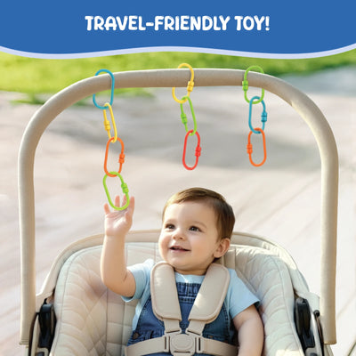 Baby seated in a padded stroller reaches upward to touch colorful plastic Linkies hanging from the stroller handle. The links in yellow, green, blue, orange, and red are clipped individually and in short chains. A blue banner at the top reads “Travel-Friendly Toy.” The background shows a softly blurred outdoor path, emphasizing on-the-go play and reach-and-grasp interaction.