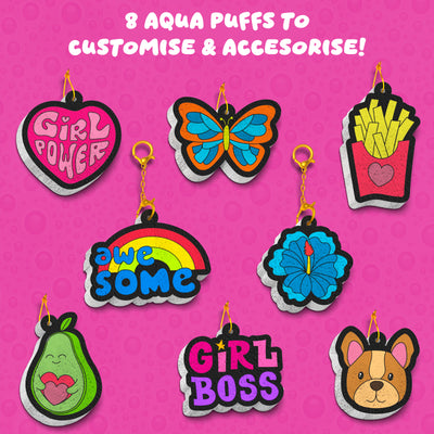 Flat lay of completed Aqua Puffs designs on bright pink background including Girl Power heart, butterfly, fries with heart, rainbow with “awesome,” blue flower, pear with heart, Girl Boss text, and dog face. Each design appears puffed with layered 3D edges and gold keychain attachments. Text states: Create 8 Aqua Puffs and attach string or keychains.