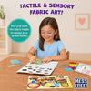 Young child seated at a table creating tactile fabric art with the Skillmatics Fab Tabs kit. Fabric pieces, picture boards, and activity materials are spread across the workspace as the child places textured fabric onto a design board, demonstrating the mess-free sensory craft experience.