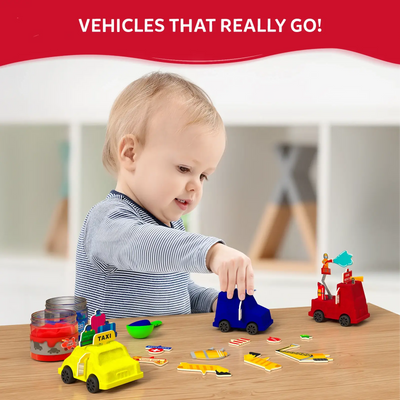 A toddler sits at a wooden table playing with bright kinetic-sand vehicles made from the Sandeezy kit, including a blue car, a yellow taxi, and a red fire engine topped with themed signs. In front of the child are scattered vehicle accessories like lights, cones, and labels. Jars of red and yellow sand sit nearby. A bold header above reads “Vehicles That Really Go!” against a red background.