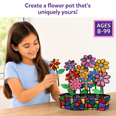 A smiling young girl in a light blue shirt places a glittery green leaf onto a colorful DIY mosaic flower while assembling a decorative flower pot craft on a wooden table. The completed arrangement features vibrant foil flowers, leaves, and a mosaic-patterned pot with an ages 8–99 badge displayed.