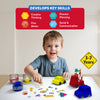 A young boy sits at a table surrounded by jars of blue, yellow, and red kinetic sand while attaching accessories to moulded sand vehicles—a yellow taxi, a red fire engine, and an unmolded blue car. Various signs and props are scattered around him. Above him, a skills panel features icons for creative thinking, process planning, fine motor skills, and social communication. A yellow road-sign badge reads “3–7 Years.”