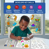 A young child sits at a table coloring an animal sticker sheet using paint markers, smiling while focused on the activity. Finished animal clings appear floating on a window behind the child. A banner at the top reads “Develops Key Skills,” with icons labeled Creative Thinking, Reflective Thinking, Fine Motor, Focus & Attention, and Spatial Reasoning, emphasizing learning benefits through hands-on play.