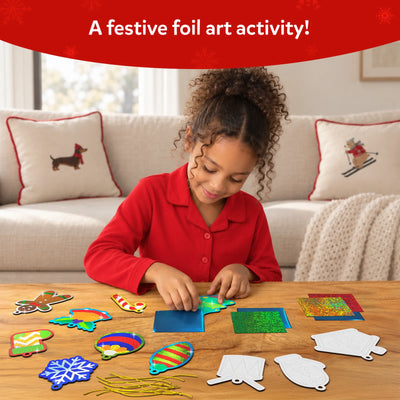 Child creating Skillmatics Foil Fun Christmas ornaments at a wooden table using colourful foil sheets and ornament templates. Finished designs including snowflakes, stockings, baubles and stars are placed around the workspace, showing a mess-free foil art activity with peel and press steps.