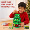 Child smiling while building a tabletop Christmas tree with Skillmatics Poke-In Art. The child holds a golden star above the decorated green fabric tree, with glitter ornaments, mini gift boxes, fabric squares and poke-in sticks on a wooden table; a blurred Christmas tree appears in the background.
