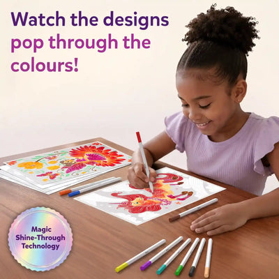 A young girl seated at a wooden table smiling while coloring Paint-Over Art sheets with markers; completed bright floral and fantasy designs visible, open kit box and scattered markers around, text reads Watch the designs pop through the colors, Magic Shine-Through badge shown.