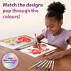 A young girl seated at a wooden table smiling while coloring Paint-Over Art sheets with markers; completed bright floral and fantasy designs visible, open kit box and scattered markers around, text reads Watch the designs pop through the colors, Magic Shine-Through badge shown.